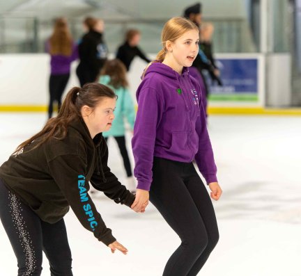 An image of two female students, dressed in casual sportswear, learning to skate at an ice skating venue